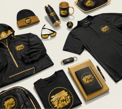 A coordinated merchandise set arranged on a light neutral background, including a black polo shirt, jacket, folded T-shirt, backpack, mug, notebook, sunglasses, key fob, and hat, all featuring a circular gold-and-black logo with the text 'The Kraft'.